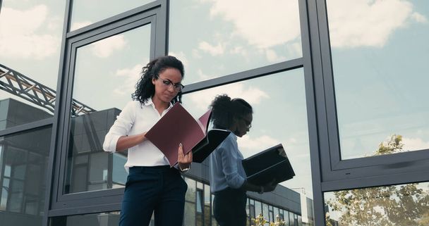 busy african american businesswoman with folders standing near building on urban street - 写真・画像
