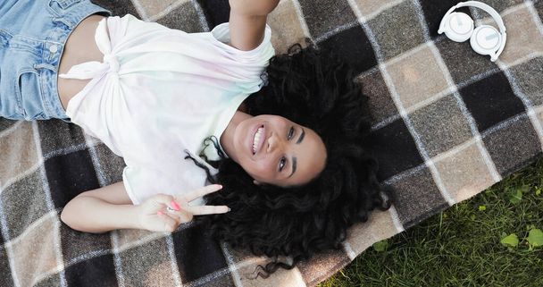 ピクニック毛布の上に横たわっている間、平和のサインを示す幸せなアフリカ系アメリカ人女性のトップビュー  - 写真・画像