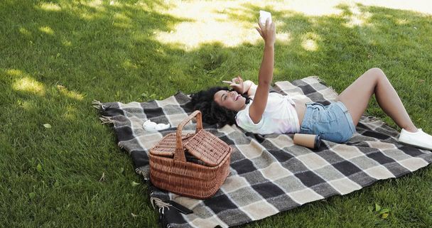 Femme afro-américaine joyeuse prenant selfie sur smartphone et couché sur couverture dans le parc  - Photo, image