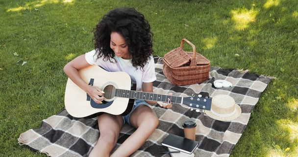overhead view of happy african american woman playing acoustic guitar during picnic in park  - Фото, изображение