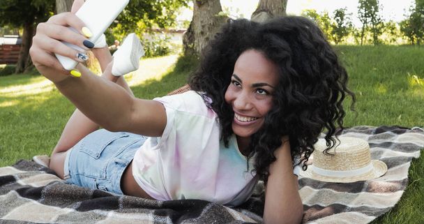 Vidám afro-amerikai nő, aki szelfit készít az okostelefonján, miközben a takarón fekszik a parkban.  - Fotó, kép