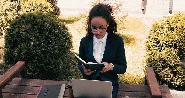 Mujer de negocios afroamericana con portátil y portátil sentado en el banco  - Foto, Imagen