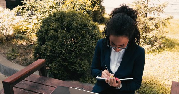 African american businesswoman writing in notebook and sitting on bench  - Φωτογραφία, εικόνα