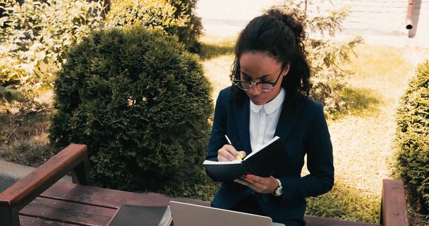 Empresaria afroamericana escribiendo en cuaderno y sentada en banco con laptop  - Foto, Imagen