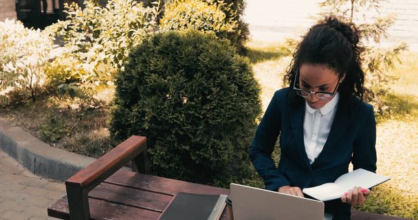 Afrykańska amerykańska bizneswoman w okularach trzyma notatnik i siedzi na ławce z laptopem  - Zdjęcie, obraz