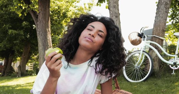 african american woman with closed eyes eating fresh apple near bicycle in park  - Zdjęcie, obraz