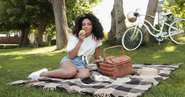 tevreden Afrikaan amerikaanse vrouw houden appel en zitten op picknick deken in park  - Foto, afbeelding