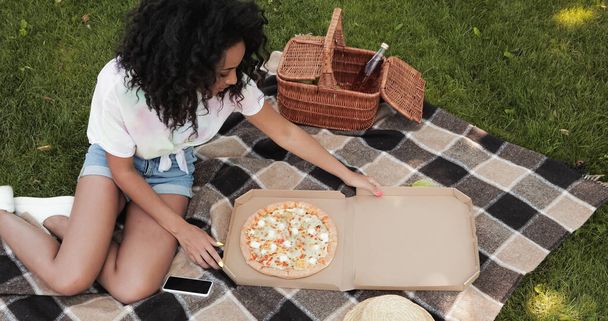 θέα υψηλής γωνίας της αφρικανικής αμερικανικής γυναίκας κοιτάζοντας πίτσα κατά τη διάρκεια πικ-νικ στο πάρκο  - Φωτογραφία, εικόνα