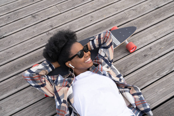 Zorgeloos Afrikaans meisje chill liggend op longboard luisteren muziek glimlachen en ontspannen. Levensstijl van stadsbewoners - Foto, afbeelding