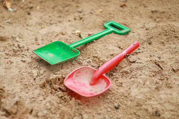 Red and green kids toddlers plastic play shovels laying on a sandbox sand background. Foto tirada em um ambiente sombrio. - Foto, Imagem