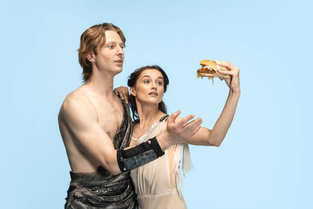 Burger and fast food. Young couple of ballet dancers in ancient Rome costums at blue studio. - Фото, зображення