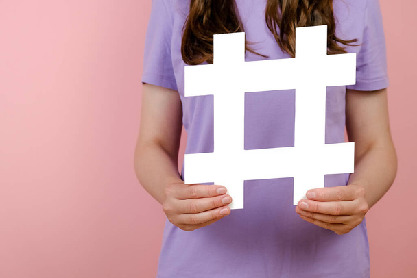 Closeup of young woman holds large big white hashtag sign, concept of trendy social media posts and blogging, viral web content, internet promotion, isolated over pink background wall in studio - Photo, image