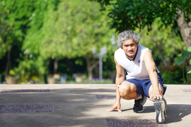 літній чоловік посміхається, бігун розігріває вправи, розтягуючи ноги в парку в хорошій атмосфері на ранковому сонці, концепція старіння суспільства, хороше здоров'я, якість життя
.                                                                          - Фото, зображення