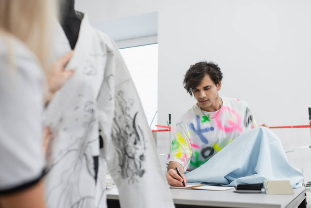 fashion designer holding tissue sample and writing in notebook near blurred colleague  - Photo, Image