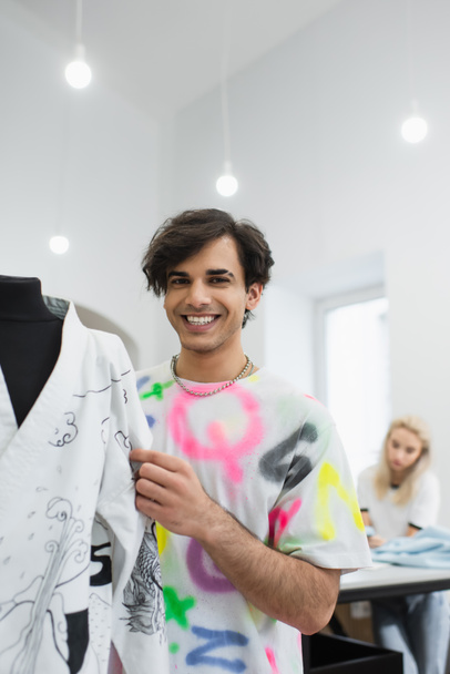 trendy designer smiling at camera near kimono on mannequin and colleague working on blurred background - Photo, Image