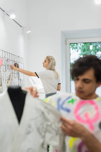 giovane designer attaccare disegni sul muro vicino collega che lavora su primo piano sfocato - Foto, immagini