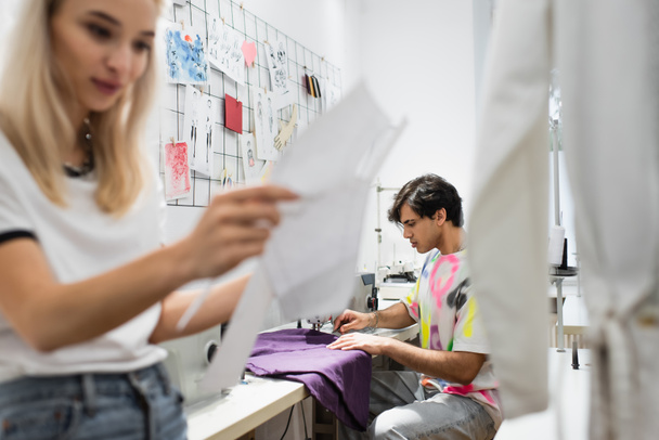 módní návrhář drží šicí vzory v blízkosti krejčí pracuje na šicím stroji - Fotografie, Obrázek