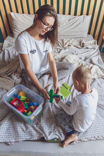 Mom and kid play together in the bedroom on a bed with plastic blocks - Zdjęcie, obraz