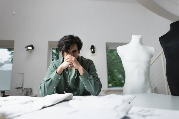 sastre reflexivo sentado cerca de patrones de costura borrosa y tejido en el taller - Foto, Imagen
