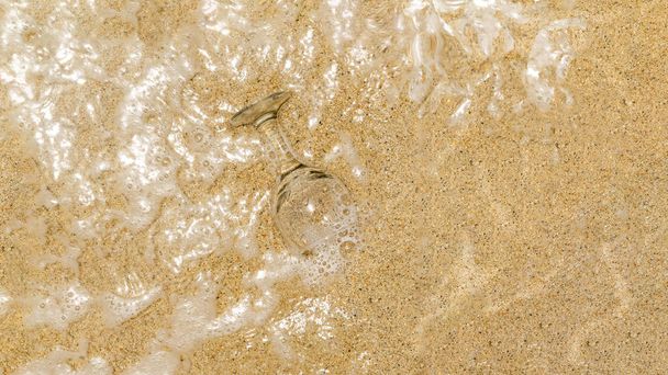 A glass forgotten on a sandy beach half covered by sea foam. Sea, sand and drinks creative concept. Minimal summer leisure design. Copy space - Fotó, kép