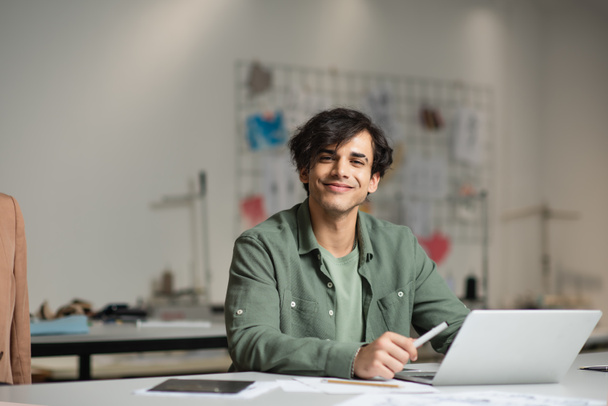 cheerful designer smiling at camera near laptop in fashion workshop - Photo, Image