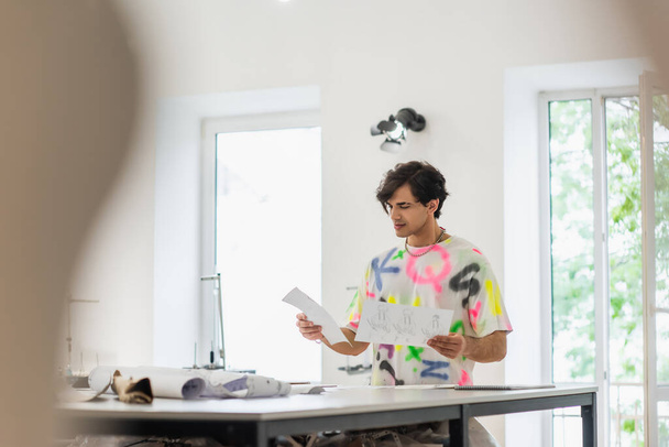 módní návrhář stojící s náčrtky na rozmazaném popředí - Fotografie, Obrázek