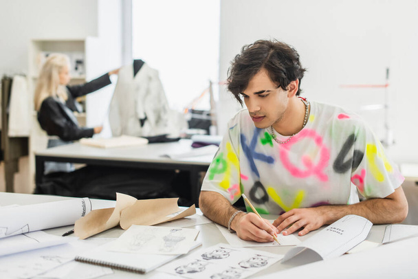 diseñador de moda creativa con lápiz mirando bocetos mientras trabaja cerca de colega borrosa en sastrería - Foto, Imagen