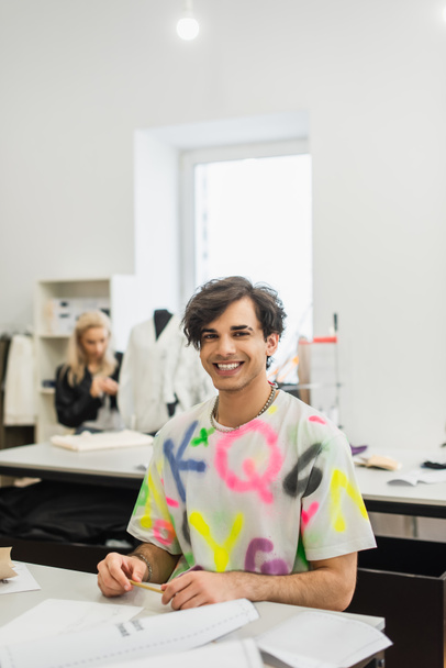 radostný módní návrhář při pohledu na kameru poblíž kolegy pracující na rozmazaném pozadí - Fotografie, Obrázek