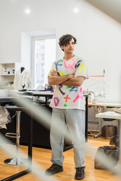 young, serious fashion designer standing with crossed arms in atelier - 写真・画像