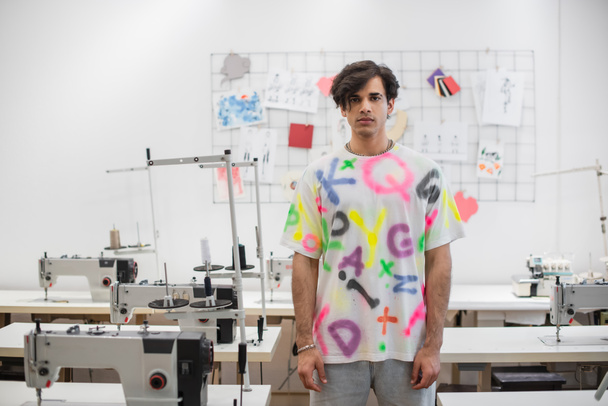 stylowy projektant mody patrząc w kamerę stojąc w pobliżu maszyn do szycia w atelier - Zdjęcie, obraz