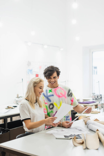 diseñadores de moda felices sosteniendo papeles mientras trabajan en sastrería - Foto, Imagen