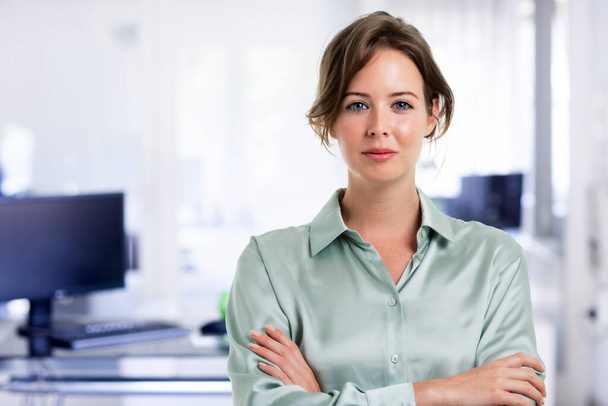 Attractive businesswoman looking at camera and smiling while standing with arms crossed at the office. - Fotoğraf, Görsel
