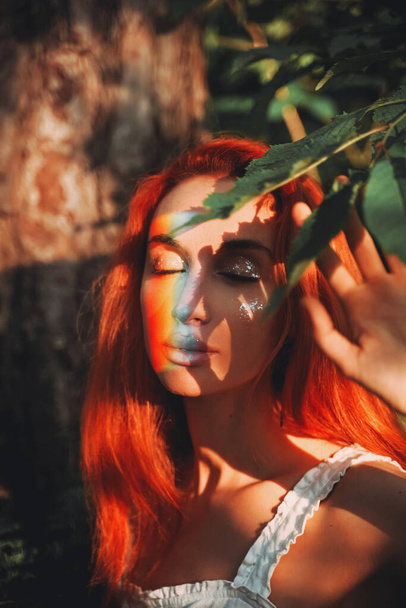 Rainbow face. Rainbow colors reflected on a beautiful redhead girl's face. Forest background - Foto, afbeelding