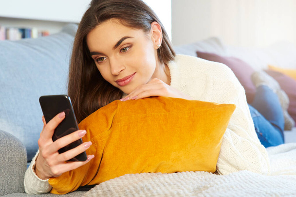 Tiro de hermosa mujer joven acostada en el sofá y mensajes de texto mientras se relaja en casa.  - Foto, Imagen
