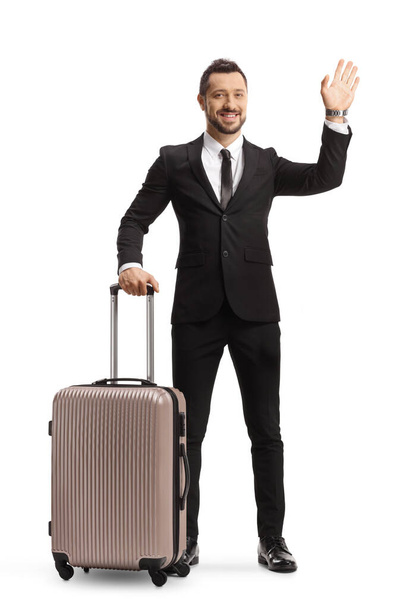Full length portrait of a young professional man in suit and tie holding a suitcase and waving isolated on white background - Photo, image
