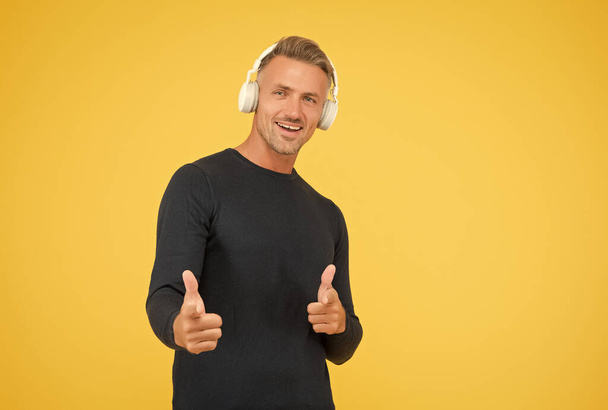 handsome cheerful man listen music in stylish headphones on yellow background pointing finger, having fun - Foto, imagen