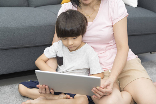 Happy Asian mother and cute little son looking and watching video in digital tablet while relaxing on couch during quarantine. - Photo, Image