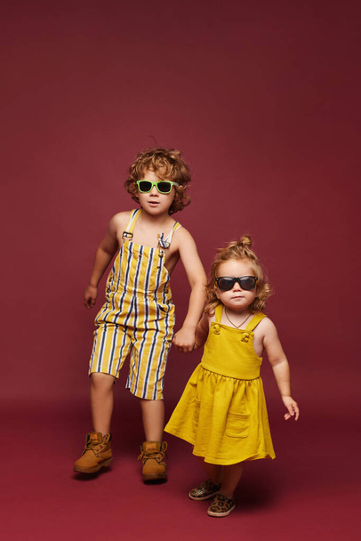 Cute little fashionable children over a pink background, isolated. Cute little girl and curly boy in summer outfits on the pink background, isolated with copy space. Kids fashion - Φωτογραφία, εικόνα