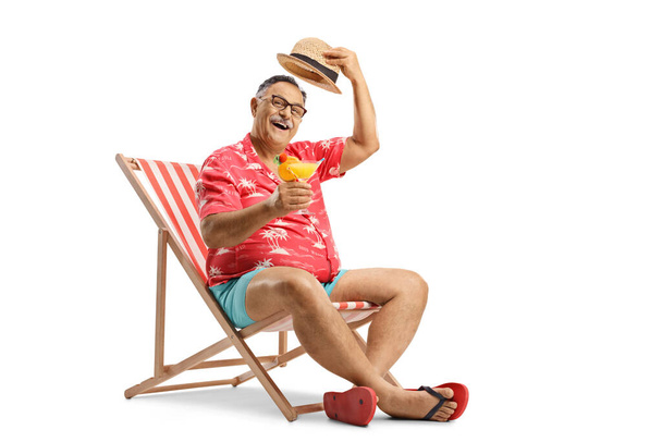 Mature tourist toasting with a cocktail, sitting on a beach chair and greeting with his hat isolated on white background - Fotografie, Obrázek