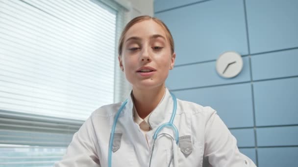 Uśmiechnięta młoda kobieta w białym płaszczu z niebieskim stetoskopem - Materiał filmowy, wideo