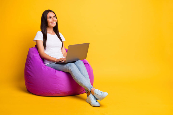 Photo of shiny young lady wear white t-shirt sitting beanbag typing modern gadget looking empty space isolated yellow color background - Foto, immagini