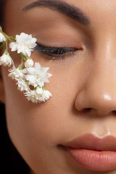 Close up half portrait of a face with beautiful skin and nude makeup, eyeliner and long eyelashes, puffy natural lips and white flowers. Fashion photography. Cosmetology and spa. Freshness of spring - Fotoğraf, Görsel