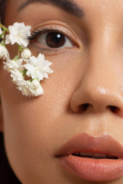 Close up half portrait of a face with beautiful skin and nude makeup, eyeliner and long eyelashes, puffy natural lips and white flowers. Fashion photography. Cosmetology and spa. Freshness of spring - Φωτογραφία, εικόνα