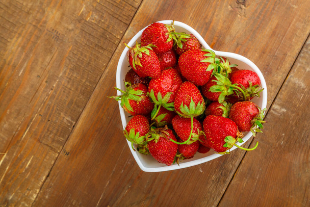Fresh large strawberries in a white plate in the shape of a heart on the table. Horizontal photo - Foto, Imagen