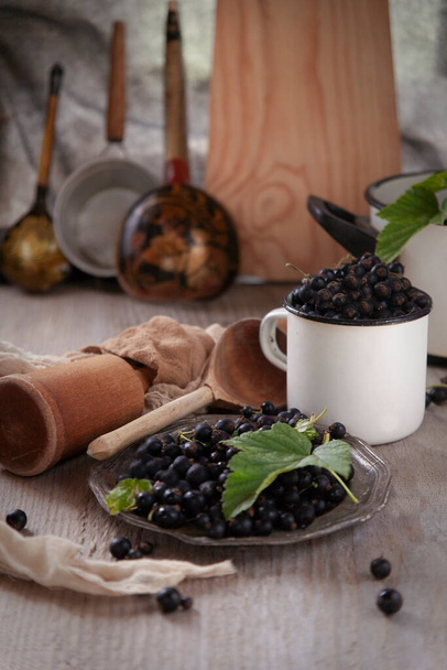 Black currant berries  in white cup and metal plate on wooden table in rustic traditional kitchen with wooden kitchen cutlery, vertical, selective focus - Fotoğraf, Görsel