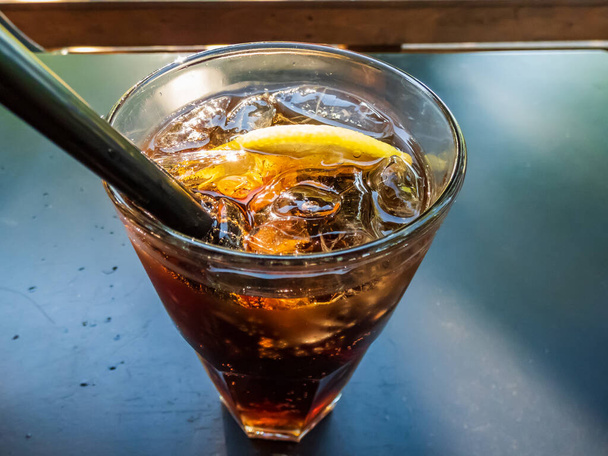 Стакан алкогольного напитка "Лонг-Айлендский чай со льдом" (водка, текила, легкий ром, тройной сек, джин и кола) с кубиками льда на тёмном столе на открытой террасе летом - Фото, изображение