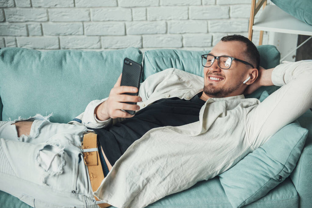 Joven hombre de negocios acostado en un sofá y sonreír mientras chatea en el móvil con auriculares - Foto, Imagen