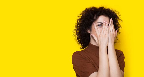 Schattige krullende vrouw bedekt haar gezicht met handpalmen glimlachend op een gele studiomuur met vrije ruimte - Foto, afbeelding