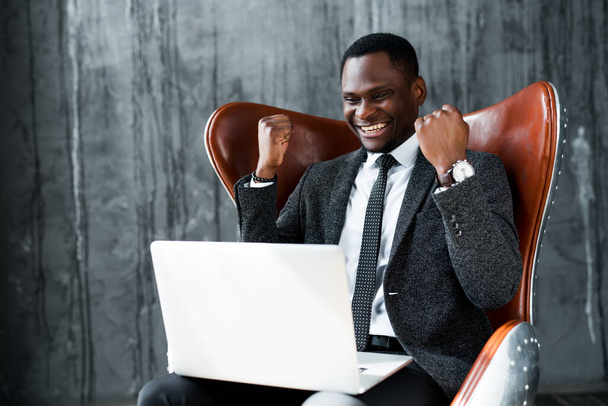 African american businessman sitting on a chair and working at a laptop rejoicing in a deal - Foto, imagen