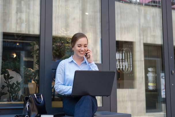 Porträt einer glücklichen jungen Geschäftsfrau, die lächelt, während sie telefoniert und mit dem Laptop arbeitet - Foto, Bild
